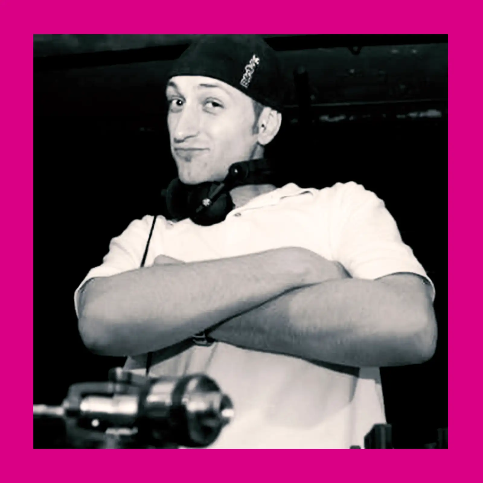 DJ posiert mit verschränkten Armen vor einem pinken Hintergrund.
